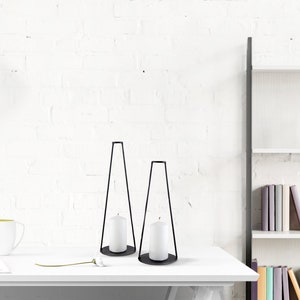 Peut inclure: Deux bougeoirs en métal noir avec des bougies blanches sur une table blanche. Les bougeoirs sont de forme triangulaire et ont une base circulaire.