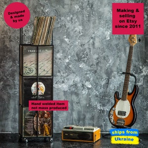 Black four-tier metal vinyl record stand on wheels, partially filled with LP albums, placed in a vintage-style room with a bass guitar and retro record player, minimalist square steel frame,