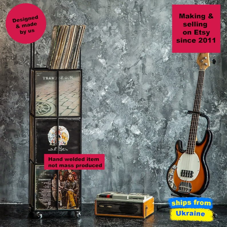 Black four-tier metal vinyl record stand on wheels, partially filled with LP albums, placed in a vintage-style room with a bass guitar and retro record player, minimalist square steel frame,