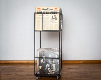 May include: A black metal record stand with two shelves. The top shelf holds a stack of vinyl records with a newspaper on top. The newspaper is titled "Good Times" and has the text "Love is all around". The bottom shelf holds a stack of vinyl records.