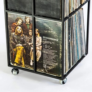 Close-up of the lower corner of a black four-tier metal vinyl record stand on wheels, showing LP albums stored vertically with visible album covers, minimalist square steel frame,