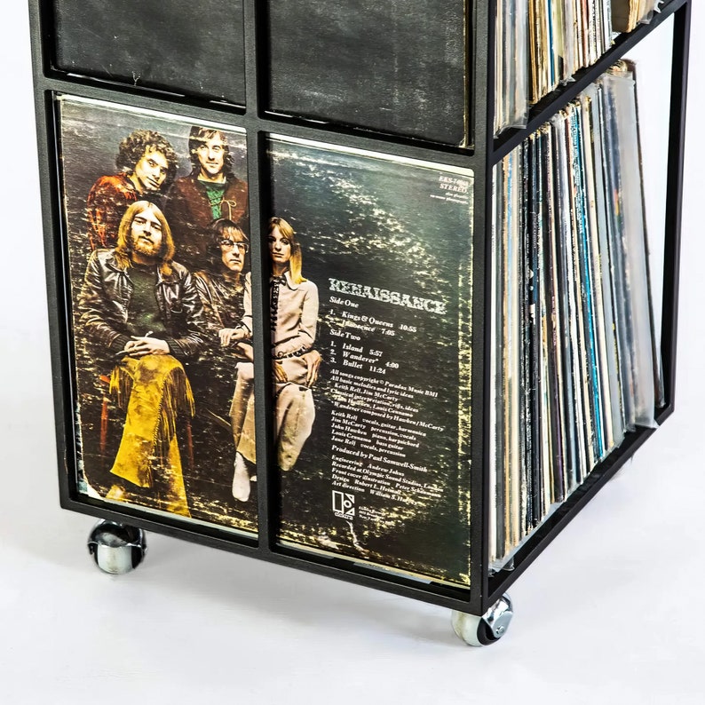 Close-up of the lower corner of a black four-tier metal vinyl record stand on wheels, showing LP albums stored vertically with visible album covers, minimalist square steel frame,