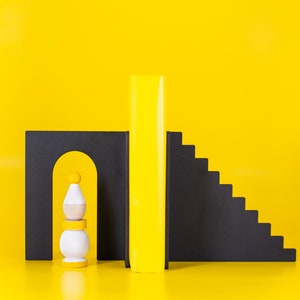 Puede incluir: Una escultura abstracta en amarillo, negro y blanco con un diseño geométrico. La escultura está hecha de madera y presenta una pieza rectangular alta, un arco curvo y un conjunto de escaleras.