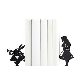 May include: Black metal bookends featuring silhouettes of Alice in Wonderland and the White Rabbit. The bookends are holding up a stack of white books.
