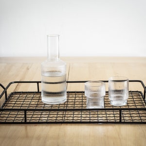 Puede incluir: Una bandeja de alambre de metal negro con una botella de vidrio transparente y dos vasos de vidrio transparentes. La bandeja está sobre una superficie de madera.