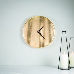 Puede incluir: Un reloj redondo dorado con agujas negras en una pared blanca, con dos candelabros de metal negro con velas blancas sobre una superficie blanca.