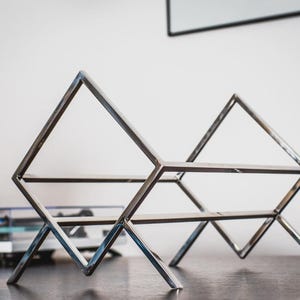 May include: A silver-colored metal record holder featuring a geometric design with two triangular side frames connected by horizontal bars. The structure is composed of thin, polished metal rods forming an open, angular frame for storing vinyl records.