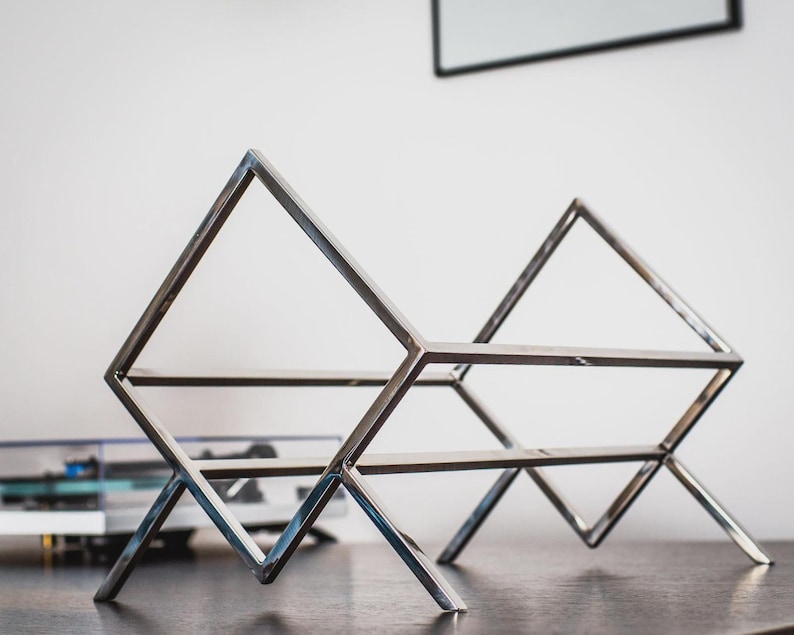 May include: A silver-colored metal record holder featuring a geometric design with two triangular side frames connected by horizontal bars. The structure is composed of thin, polished metal rods forming an open, angular frame for storing vinyl records.