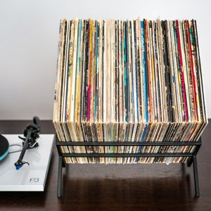 May include: A black metal wire rack holding a large collection of vinyl records in square cardboard sleeves, arranged vertically. To the left, a white turntable with a black tonearm and a blue cartridge is visible, featuring the text P3 on its surface.