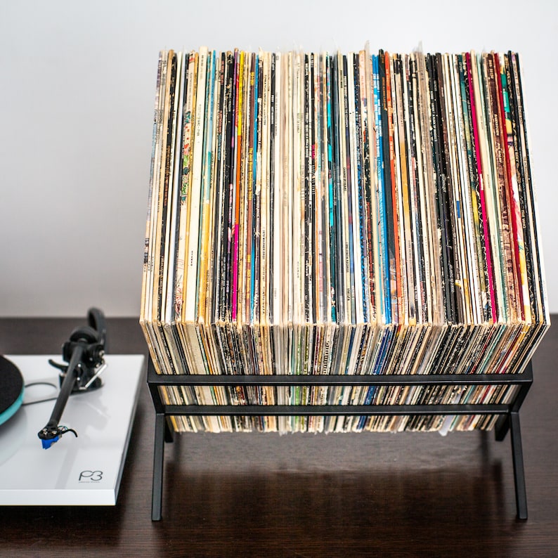 May include: A black metal wire rack holding a large collection of vinyl records in square cardboard sleeves, arranged vertically. To the left, a white turntable with a black tonearm and a blue cartridge is visible, featuring the text P3 on its surface.