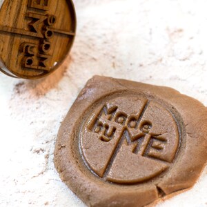 May include: A close-up of a brown cookie dough with a wooden stamp pressed into it. The stamp reads "Made by Me".