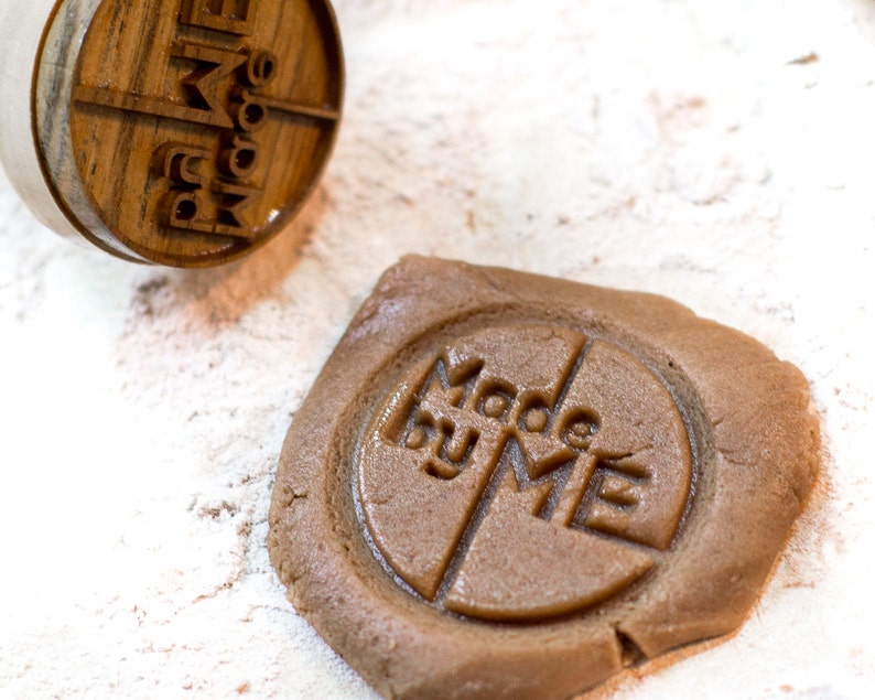 May include: A close-up of a brown cookie dough with a wooden stamp pressed into it. The stamp reads "Made by Me".