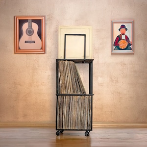May include: A black metal record storage unit with a wooden top and two shelves. The unit is filled with vinyl records and is on wheels. There are two framed pictures on the wall behind the unit, one with a guitar and the other with a person playing a banjo.