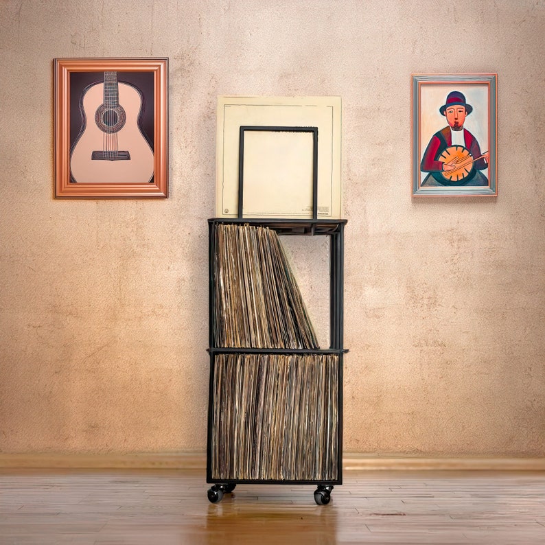 May include: A black metal record storage unit with a wooden top and two shelves. The unit is filled with vinyl records and is on wheels. There are two framed pictures on the wall behind the unit, one with a guitar and the other with a person playing a banjo.