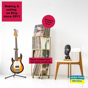Gold four-tier metal vinyl record stand on wheels, filled with LP albums, placed in a modern room with a bass guitar and designer chair, minimalist square steel frame, hand-welded in Ukraine.