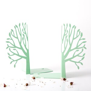 May include: A pair of green metal bookends shaped like trees with a leafy design. The bookends are on a white surface with white and pink flower petals scattered around them.