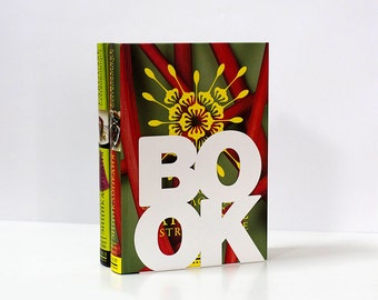 Puede incluir: Un sujetalibros blanco con forma de la palabra "BOOK" con un diseño floral verde, rojo y amarillo en la parte posterior.