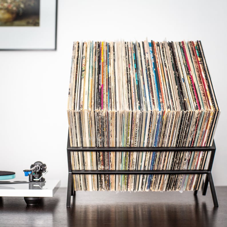 May include: A black metal vinyl record storage rack holding a large collection of vinyl records with various colorful album spines. The rack features a slanted design to display the records, positioned next to a white turntable component.