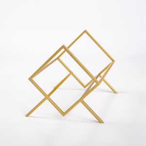 May include: A gold-colored metal rack featuring a geometric design with two interconnected square frames supported by angled legs. The structure is composed of thin, straight metal bars forming an open, angular silhouette.