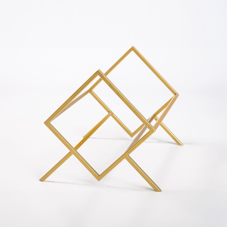 May include: A gold-colored metal rack featuring a geometric design with two interconnected square frames supported by angled legs. The structure is composed of thin, straight metal bars forming an open, angular silhouette.