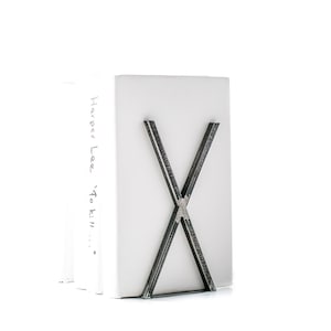 May include: A silver metal bookend with a criss-cross design holds a stack of books. The top book has the text "Harper Lee 'To Kill..." written on the spine.
