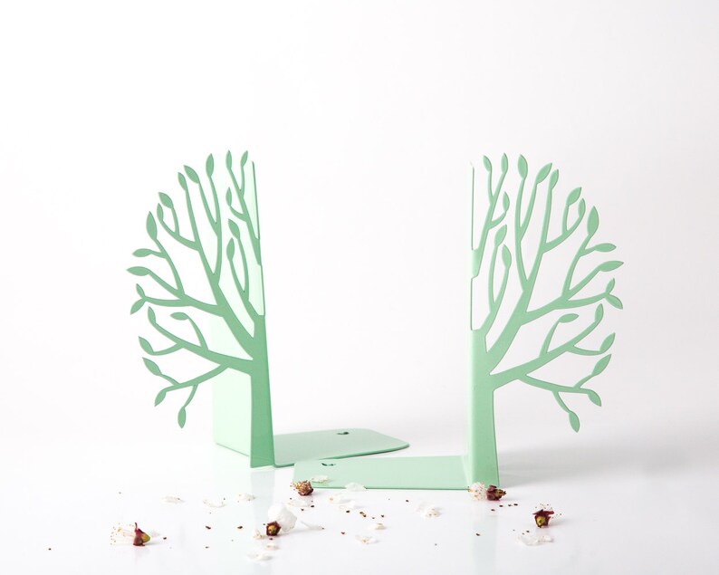 May include: A pair of green metal bookends shaped like trees. The bookends are on a white surface with white flower petals scattered around them.