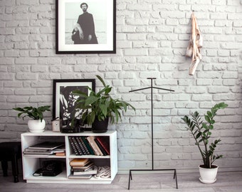 May include: White bookshelf with four shelves, filled with books and a black rotary phone. A black and white framed photo hangs above the shelf. A black metal clothing rack stands in the background against a white brick wall. Two potted plants are on the shelf.