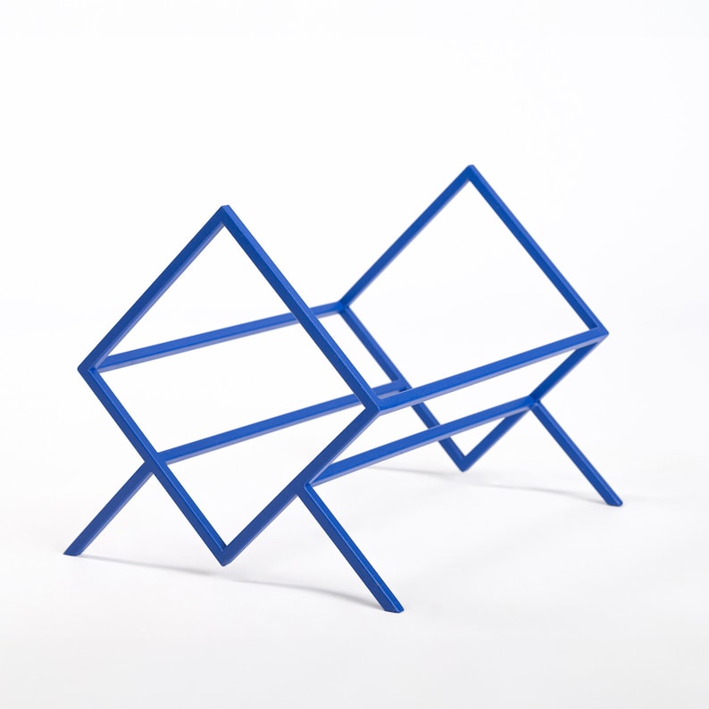 May include: A bright blue metal rack featuring a geometric, open-frame design with two diamond-shaped side supports connected by horizontal bars, creating a cradle-like structure with four angled legs for stability and storage.