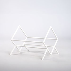 May include: A white metal rack featuring a geometric frame with two triangular side panels connected by horizontal bars. The structure is composed of thin, straight metal rods arranged in an open, angular design with four legs.