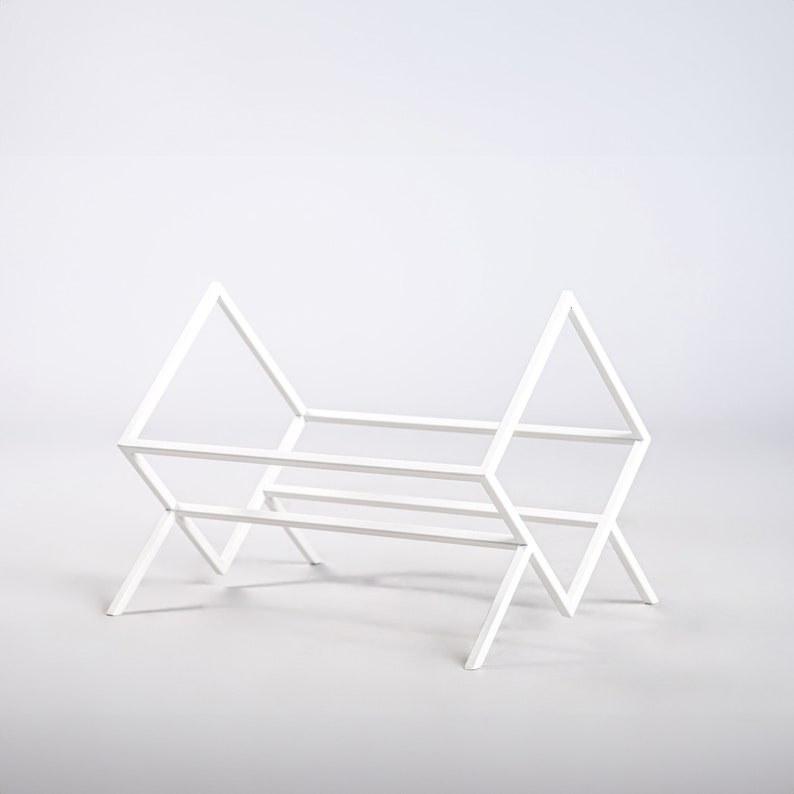 May include: A white metal rack featuring a geometric frame with two triangular side panels connected by horizontal bars. The structure is composed of thin, straight metal rods arranged in an open, angular design with four legs.