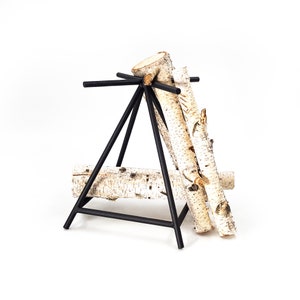 May include: Black metal log rack with three birch logs stacked on top. The rack is a triangular shape with a crossbar at the top.