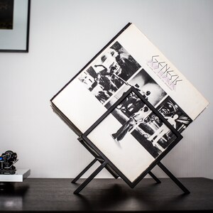 May include: A black metal wire record stand holding a vinyl album titled The Lamb Lies Down on Broadway by Genesis. The album cover features a black and white collage of photographs. The stand is angled to display the record cover prominently.