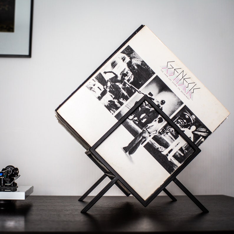 May include: A black metal wire record stand holding a vinyl album titled The Lamb Lies Down on Broadway by Genesis. The album cover features a black and white collage of photographs. The stand is angled to display the record cover prominently.