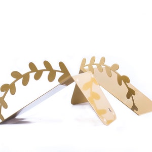 May include: Two gold metal bookends shaped like a leafy branch. The bookends are angled to create a V-shape.