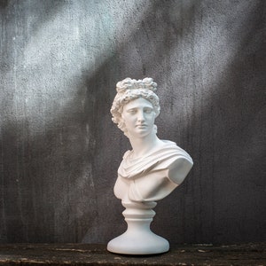 Könnte beinhalten: Weiße Gipsbüste einer Frau mit einer Blätterkrone auf dem Kopf. Die Büste steht auf einem weißen Sockel.