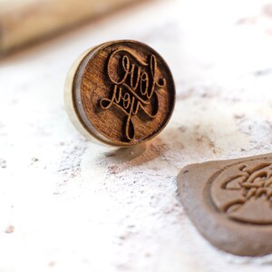 May include: A wooden stamp with the words "Love you" carved into it. The stamp is on a white surface with a brown cookie that has been stamped with the same design.