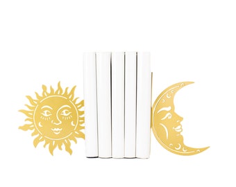 May include: Gold metal bookends shaped like a sun and a crescent moon. The sun and moon have faces with closed eyes and a smile. The bookends are holding up a stack of white books.