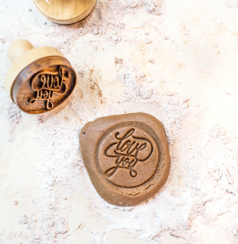 May include: A brown cookie with the words "Love You" imprinted on it. The cookie is sitting on a light brown surface.