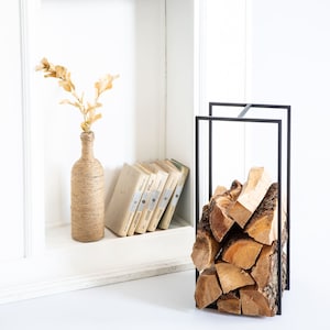 May include: Black metal firewood rack with stacked firewood logs. The rack is in a white room with a vase of dried flowers and books on a shelf.