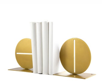 Puede incluir: Juego de sujetalibros de metal dorado con un diseño moderno. Cada sujetalibros presenta una forma circular con una ranura vertical u horizontal y una base rectangular. Libros blancos se sostienen entre los sujetalibros.