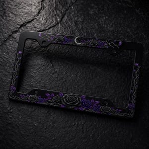 May include: A rectangular black license plate frame featuring a decorative design of dark purple and black roses with thorny vines. A small silver crescent moon is positioned at the top center of the frame, surrounded by the floral pattern.