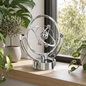 May include: A kinetic sculpture featuring a series of polished silver-colored metal rings and spheres arranged in a rotating orbital design. The object is mounted on a circular base and consists of multiple interconnected rings that surround a central sphere.