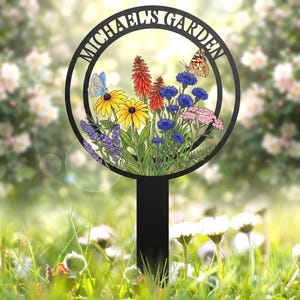 Puede incluir: Un estaca de jardín circular de metal negro con el texto MICHAEL'S GARDEN arqueado en la parte superior. El centro muestra una ilustración colorida de equináceas amarillas, kniphofia rojas, acianos azules y dos mariposas.