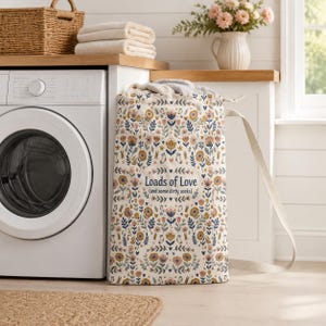 May include: A tall, off-white fabric laundry bag with a repeating pattern of colorful flowers, leaves, and small socks. The center features the text "Loads of Love (and some dirty socks)" in a dark blue font, with two sturdy canvas shoulder straps.
