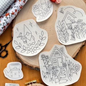 May include: Five embroidery patterns on white fabric featuring a gnome, a birdhouse, a house, a teapot, and a teacup. Each pattern is surrounded by flowers and leaves.