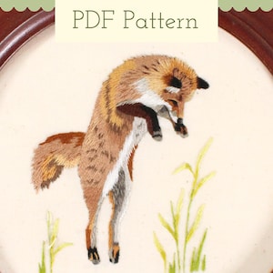 以下が含まれることがあります： 緑の草のフィールドでジャンプしている赤いキツネの刺繍画像。キツネは茶色と白の毛皮で詳細に描かれています。画像は茶色の木製の円形枠で囲まれています。