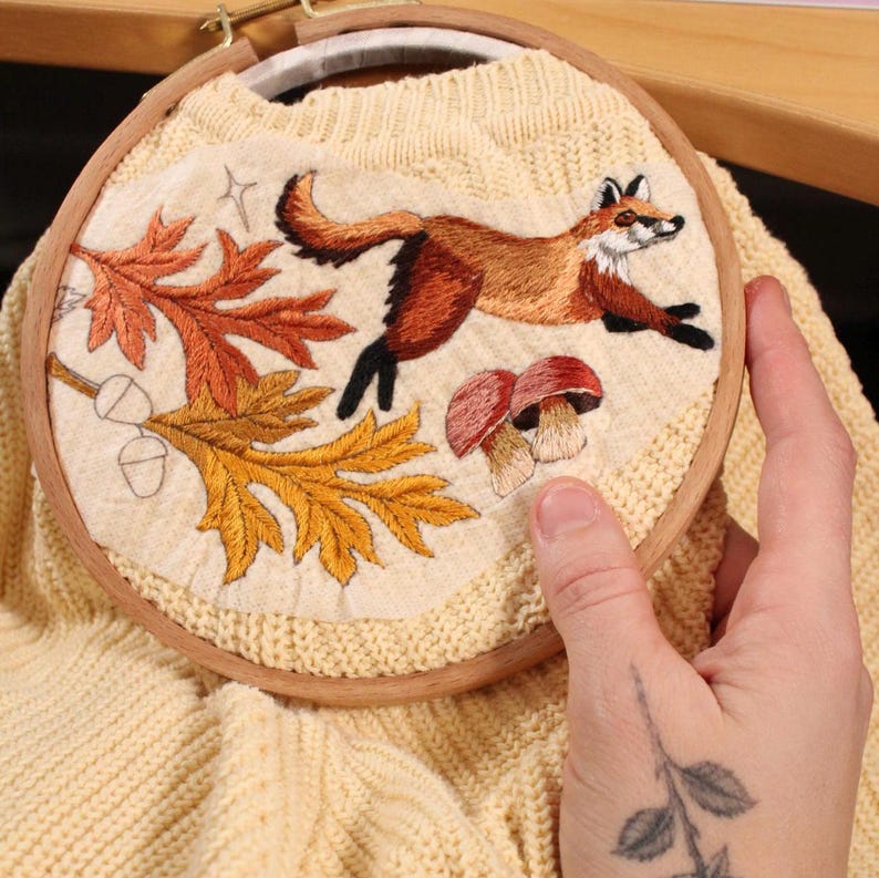 hand holding in progress embroidery of fox, mushrooms, and leaves