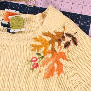 close up of a yellow knit sweater with embroidery along the neckline. The embroidery features colorful leaves, rose hips, and acorns