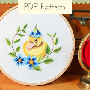 以下が含まれることがあります： 青い帽子をかぶった黄色いアヒルの刺繍アート。青い花と緑の葉で囲まれています。「PDF Pattern」の文字が上部に表示されています。刺繍は木製の枠にセットされています。
