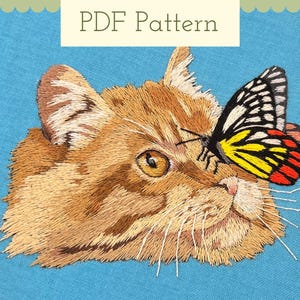 Puede incluir: Imagen bordada de un gato jengibre con una mariposa amarilla y negra en la nariz. El gato mira hacia la derecha y la mariposa hacia la izquierda. El fondo es una tela azul claro.